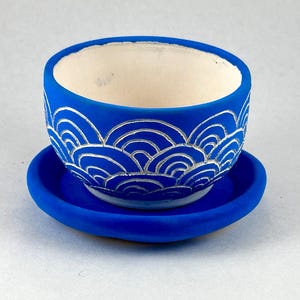 May include: A small, round, blue ceramic planter with a matching saucer. The planter features a wave-like pattern in silver around the exterior. The interior of the planter is white.