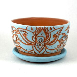 May include: A handcrafted ceramic planter with a light blue exterior and a terracotta interior. The exterior features a detailed, raised floral design in a contrasting terracotta color. The planter sits on a matching light blue saucer.