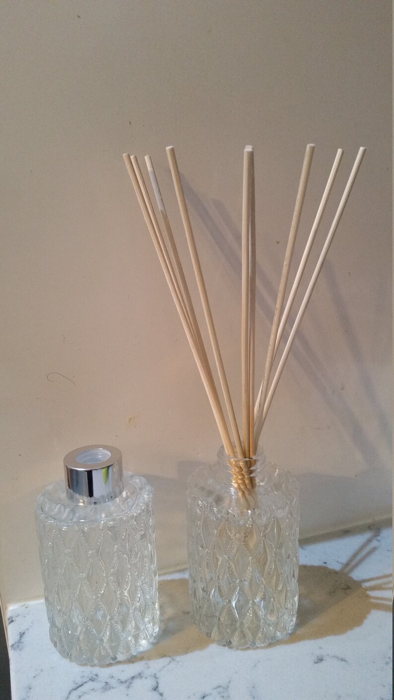 May include: Two clear glass bottles with textured surfaces, one filled with a liquid and the other with wooden sticks. The bottles have silver caps.