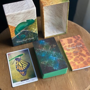 Puede incluir: Una baraja de cartas de or&aacute;culo con un patr&oacute;n de piel de serpiente verde, azul y marr&oacute;n en la parte delantera. Las cartas est&aacute;n en una caja con un patr&oacute;n similar. La caja est&aacute; abierta y las cartas son visibles. Hay una gu&iacute;a con un patr&oacute;n similar en la parte delantera. La gu&iacute;a est&aacute; cerrada. Las cartas se titulan "Familiar Field Oracle".