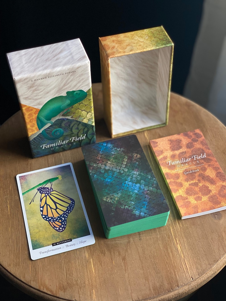 Puede incluir: Una baraja de cartas de or&aacute;culo con un patr&oacute;n de piel de serpiente verde, azul y marr&oacute;n en la parte delantera. Las cartas est&aacute;n en una caja con un patr&oacute;n similar. La caja est&aacute; abierta y las cartas son visibles. Hay una gu&iacute;a con un patr&oacute;n similar en la parte delantera. La gu&iacute;a est&aacute; cerrada. Las cartas se titulan "Familiar Field Oracle".