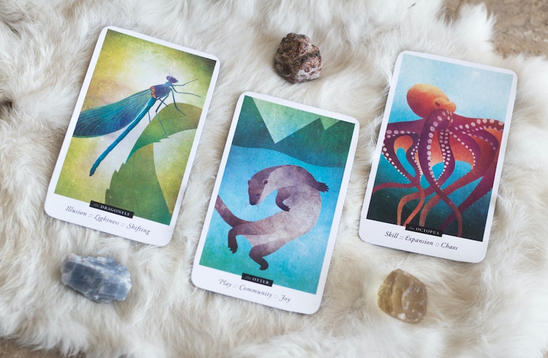 Puede incluir: Tres cartas de una baraja de cartas de or&aacute;culo animal. La primera carta presenta una lib&eacute;lula azul con el texto "Lib&eacute;lula: Ilusi&oacute;n - Ligereza - Cambio". La segunda carta presenta una nutria marr&oacute;n nadando con el texto "Nutria: Juego - Comunidad - Alegr&iacute;a". La tercera carta presenta un pulpo rojo con el texto "Pulpo: Habilidad - Expansi&oacute;n - Caos".