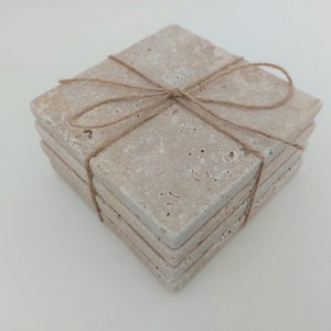 Op de afbeelding: Set van vier beige stenen onderzetters die met touw aan elkaar zijn gebonden. De onderzetters zijn vierkant en hebben een ruw, getextureerd oppervlak.