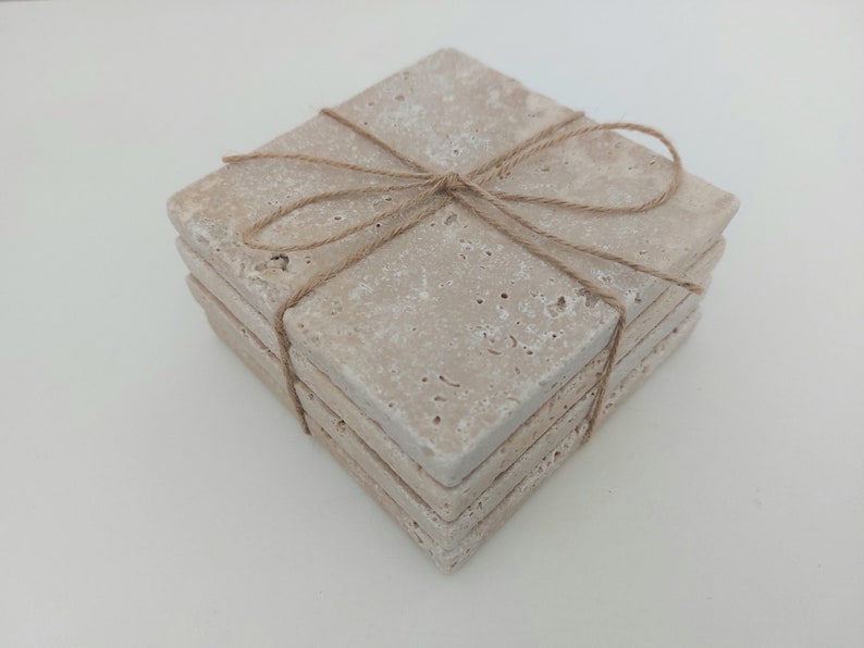Op de afbeelding: Set van vier beige stenen onderzetters die met touw aan elkaar zijn gebonden. De onderzetters zijn vierkant en hebben een ruw, getextureerd oppervlak.