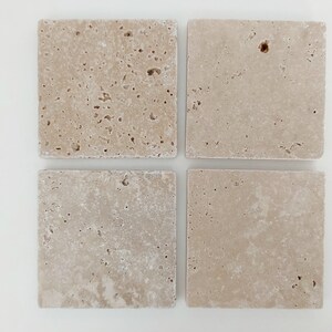 Op de afbeelding: Vier vierkante beige travertijn tegels met een ruw, getextureerd oppervlak.