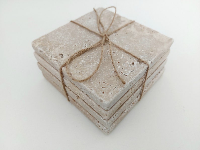 Op de afbeelding: Set van vier beige stenen onderzetters gebonden met touw. De onderzetters zijn vierkant en hebben een ruw, getextureerd oppervlak.