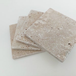 Op de afbeelding: Set van vier beige travertijnstenen onderzetters. De onderzetters zijn vierkant en hebben een ruw, getextureerd oppervlak.