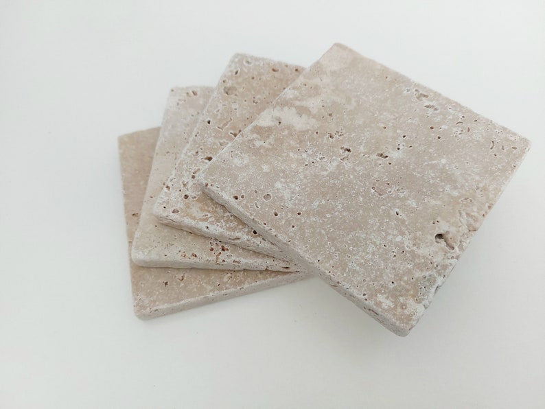 Op de afbeelding: Set van vier beige travertijnstenen onderzetters. De onderzetters zijn vierkant en hebben een ruw, getextureerd oppervlak.