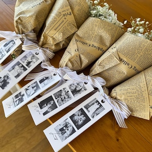 May include: Several bouquets of white flowers wrapped in vintage newspaper. Each bouquet is tied with a striped ribbon and a tag with black and white photos. The bouquets are arranged on a wooden surface.