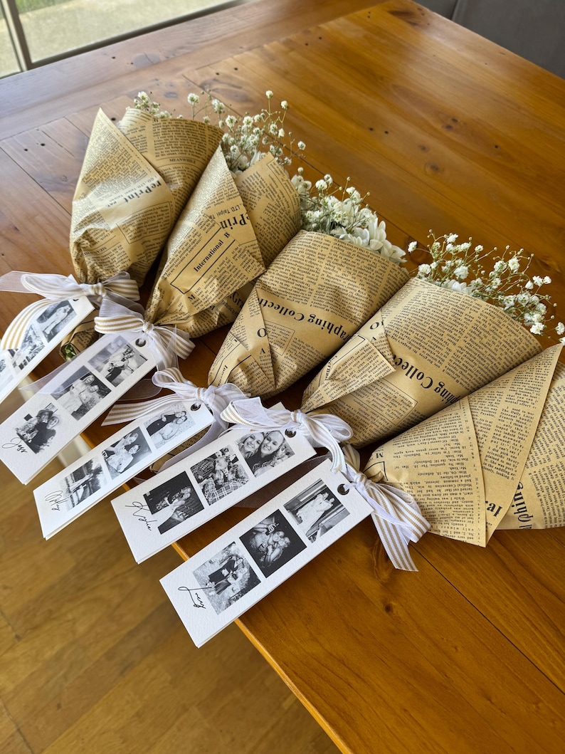 May include: Several bouquets of white flowers wrapped in vintage newspaper. Each bouquet is tied with a striped ribbon and a tag with black and white photos. The bouquets are arranged on a wooden surface.