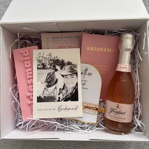 May include: A pink gift box filled with white tissue paper. The box contains a pink card with the text "Will you be my Bridesmaid?" and a photo of a woman wearing a cowboy hat. There is also a pink card with the text "Heather" and a bottle of Freixenet Italian Rose sparkling wine.