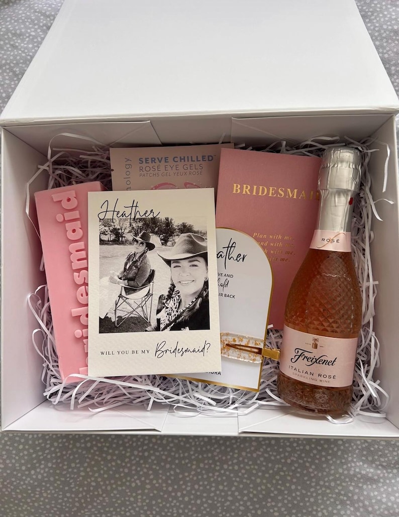 May include: A pink gift box filled with white tissue paper. The box contains a pink card with the text "Will you be my Bridesmaid?" and a photo of a woman wearing a cowboy hat. There is also a pink card with the text "Heather" and a bottle of Freixenet Italian Rose sparkling wine.