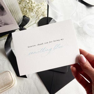 May include: A white card with the text "Lauren, thank you for being my something blue" in black and light blue script. The card is held over a black envelope, with a small jewelry box and black ribbon in the background.