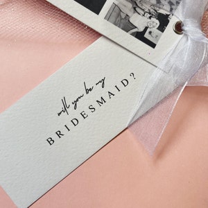 Bookmark style photo card strip in black and white. Features 3 personalised photos and name on the front. The back says will you be my bridesmaid or maid of honour.Tied with a white organa bow.