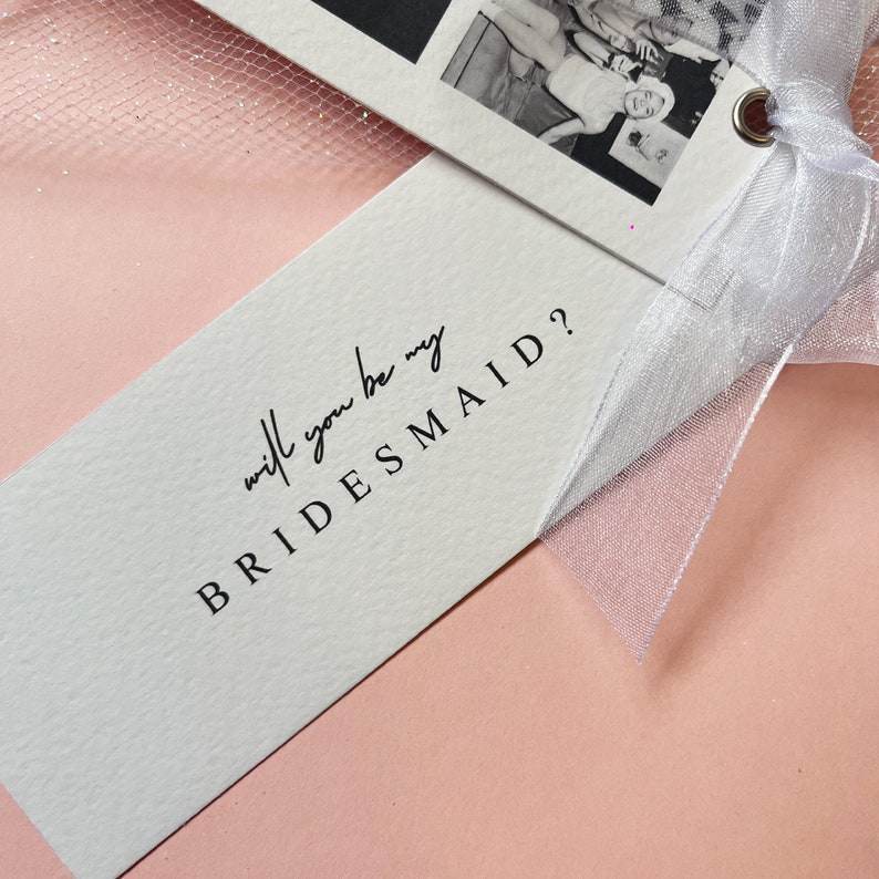 Bookmark style photo card strip in black and white. Features 3 personalised photos and name on the front. The back says will you be my bridesmaid or maid of honour.Tied with a white organa bow.