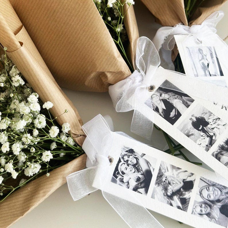 May include: Bouquets of white baby's breath flowers wrapped in brown paper. Each bouquet is adorned with a white ribbon and a tag featuring black and white photographs. The tags are attached with a small metal fastener.