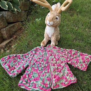 May include: A handmade, open-front crochet cardigan in shades of pink and green, with a textured, raised stitch pattern. A plush, tan-colored rabbit toy with long ears sits behind the cardigan on a grassy surface.
