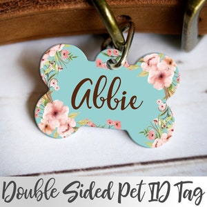 May include: A light blue bone-shaped pet ID tag with a floral design and the name "Abbie" printed in brown text. The tag is double-sided.