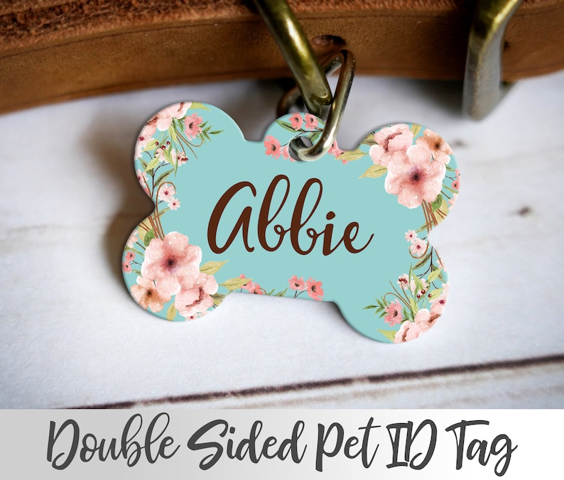 May include: A light blue bone-shaped pet ID tag with a floral design and the name "Abbie" printed in brown text. The tag is double-sided.