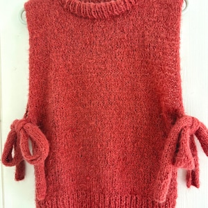 Può includere: Un top senza maniche in maglia color ruggine, con scollatura rotonda e dettagli a fiocco sulle spalle. Il top è realizzato in un filato strutturato ed è appeso a una gruccia verde chiaro su sfondo bianco.