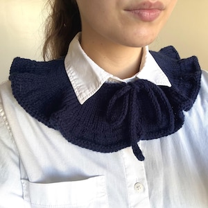 Peut inclure: Un col amovible en tricot bleu marine foncé doté d'un bord à volants et d'une fermeture à nouer au centre. L'accessoire est porté sur une chemise blanche à col boutonné, mettant en valeur un motif en tricot texturé et une forme arrondie.