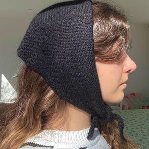 May include: A black knitted headscarf tied under the chin. The head covering is made of a dark, textured material and is worn over the hair. The scarf is tied with a knot, and the ends hang down. The person is wearing a striped shirt.