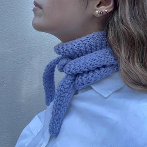 May include: A light periwinkle blue knitted neck scarf featuring a textured garter stitch pattern. The scarf is tied in a simple knot at the front, with two tapered ends hanging down. It is worn over a crisp white collared button-down shirt.