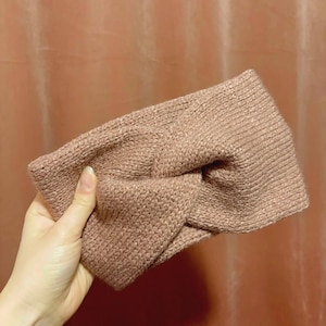 Peut inclure: Un bandeau tricoté de couleur rose clair. Le bandeau présente un motif de nœud torsadé au centre. La matière semble être un tricot doux et texturé. Le bandeau est tenu par une main.