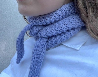 May include: A light purple knitted scarf with a textured stitch pattern, wrapped around the neck and tied in a simple knot at the front. The scarf is made of thick yarn and features two long, tapered ends that hang down from the central knot.