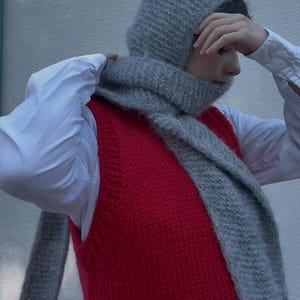 May include: A person wearing a red knitted vest over a white shirt, with a grey knitted hood and scarf. The scarf is wrapped around the neck, and the hood covers the head. The outfit is a cosy, winter-themed ensemble.