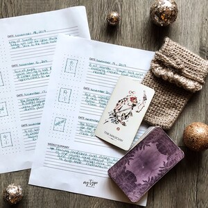 May include: A tarot card reading journal with handwritten notes and dates. The journal is open to a page with the text "The Magician" and a drawing of a person holding a wand. The journal is on a wooden surface with a knitted brown mitten and gold ornaments.