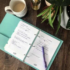 May include: A teal planner with a white interior and a purple pen. The planner has a section for morning and bedtime routines. The morning section includes a list of things to be grateful for and intentions for the day. The bedtime section includes a list of things that made the day enjoyable.