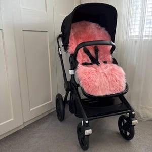 May include: A black pushchair featuring a pink, fluffy seat liner. The pushchair has a black frame, wheels, and hood. The seat liner is a bright pink colour and looks soft and plush.