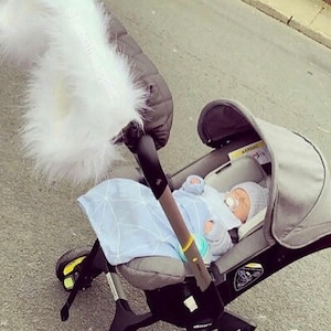 May include: A grey stroller with a white fluffy handle cover and a baby inside wearing a blue and white outfit. The stroller has a grey canopy and a black handle.