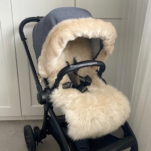 Può includere: Un rivestimento per passeggino in pelliccia sintetica color crema con un cappuccio abbinato. Il rivestimento è in un passeggino con telaio nero e capottina in tessuto grigio. Il testo "@furcoatnonappies_" è visibile sulla maniglia.