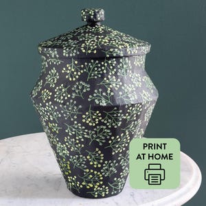 Pode incluir: Um pote preto com tampa apresentando um padrão repetitivo de ramos verde-claros e brancos. O pote tem um corpo angular facetado e uma tampa correspondente com um pequeno puxador. Uma etiqueta quadrada verde-clara contém o texto PRINT AT HOME.