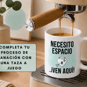May include: A white coffee mug with the text "NECESITO ESPACIO ...&iexcl;VEN AQU&Iacute;!" and a heart-shaped lock and key design. Coffee is being poured from an espresso machine. A sign reads "COMPLETA TU PROCESO DE SANACI&Oacute;N CON UNA TAZA A JUEGO."