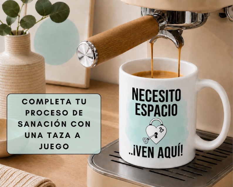 May include: A white coffee mug with the text "NECESITO ESPACIO ...&iexcl;VEN AQU&Iacute;!" and a heart-shaped lock and key design. Coffee is being poured from an espresso machine. A sign reads "COMPLETA TU PROCESO DE SANACI&Oacute;N CON UNA TAZA A JUEGO."