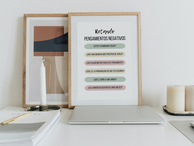 A framed print with the title "Retando Pensamientos Negativos" and a list of questions in Spanish to challenge negative thoughts. The print is on a white background with pastel colored boxes containing the questions. The print is framed in a light wood frame and is sitting on a desk with a laptop, candles, and a book.