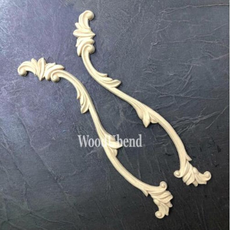 May include: Two cream-colored, curved wooden decorative appliques with a floral and leaf design. The appliques are symmetrical, with a scroll-like top and a leaf-shaped bottom. The text "Wood bend" is printed on the bottom left applique.