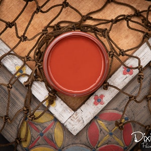 K&ouml;nnte beinhalten: Ein kleines Glas mit roter Farbe mit gl&auml;nzendem Finish. Das Glas steht auf einer Holzoberfl&auml;che mit einem braunen Seilnetz.