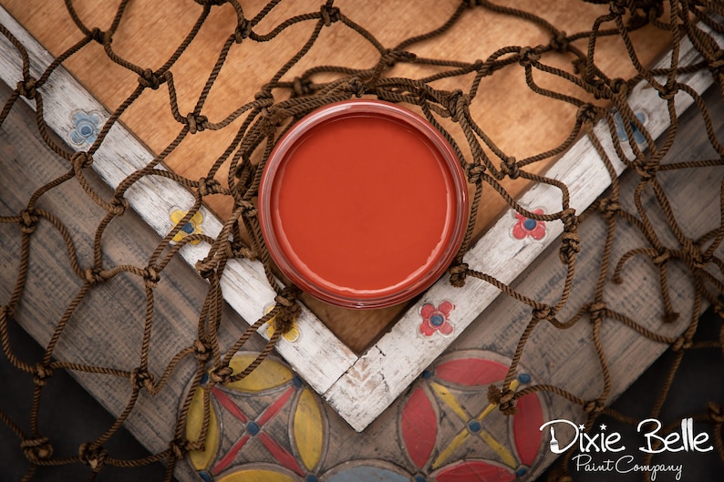 K&ouml;nnte beinhalten: Ein kleines Glas mit roter Farbe mit gl&auml;nzendem Finish. Das Glas steht auf einer Holzoberfl&auml;che mit einem braunen Seilnetz.