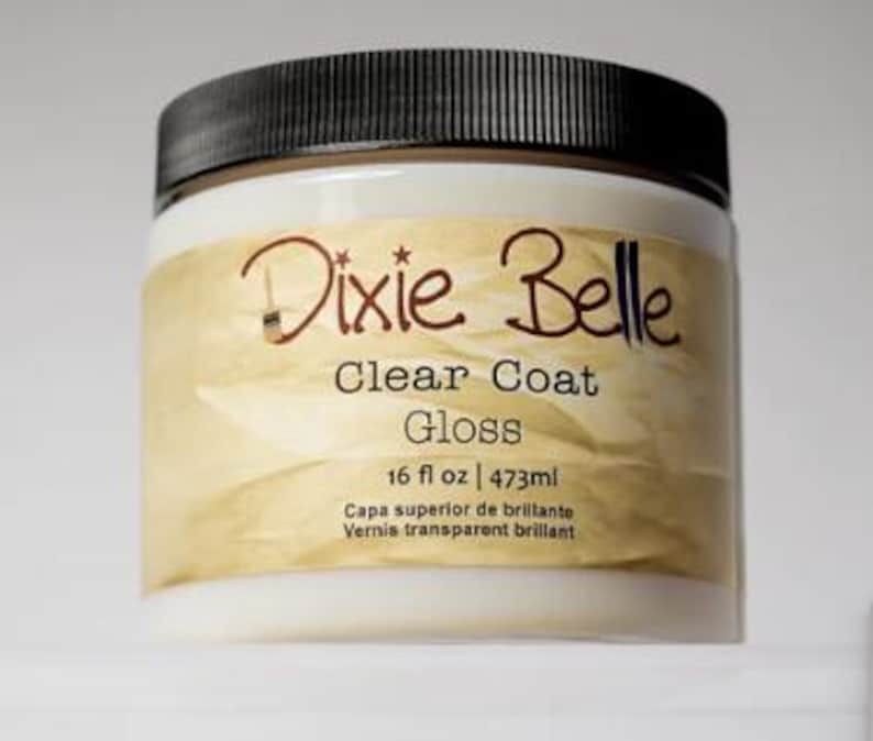 Może przedstawiać: Okrągły plastikowy pojemnik z czarną zakrętką zawierający lakier Dixie Belle Clear Coat Gloss. Beżowa etykieta zawiera nazwę marki, rodzaj produktu oraz objętość 473 ml wydrukowaną czarnym tekstem na teksturowanym tle.