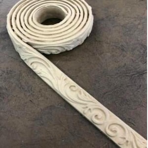 May include: A roll of off-white decorative molding with an ornate, repeating floral pattern. The molding is coiled, with a portion unrolled, revealing the detailed design. The background is a dark, textured surface.