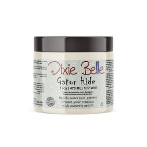 May include: A white jar with a black lid labeled "Dixie Belle Gator Hide 16oz | 473 ML | Stir Well". The label also says "Capa transparente extra duradera Vernis transparent extra durable Repels water (not gators) Protect your creation with nature's armor".