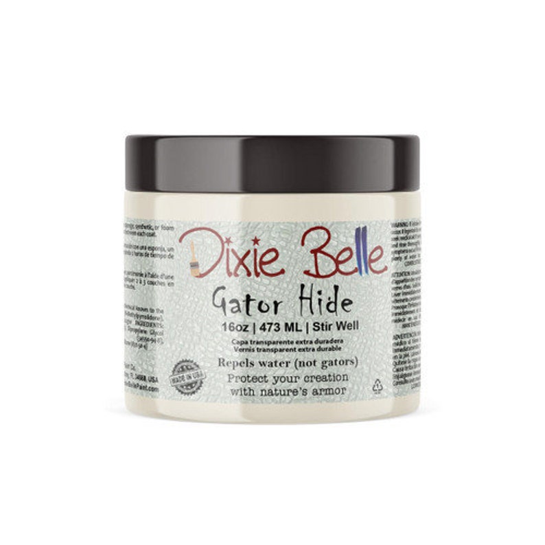 May include: A white jar with a black lid labeled "Dixie Belle Gator Hide 16oz | 473 ML | Stir Well". The label also says "Capa transparente extra duradera Vernis transparent extra durable Repels water (not gators) Protect your creation with nature's armor".