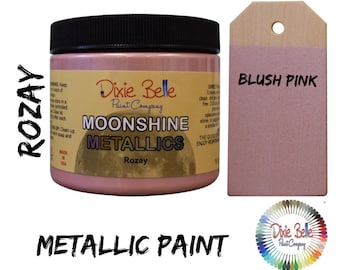 May include: A jar of Dixie Belle Moonshine Metallic paint in the color Rozay, a blush pink. The jar is labeled with the Dixie Belle logo and the words "Moonshine Metallics" and "Rozay". A wooden tag with the words "Blush Pink" is shown next to the jar.