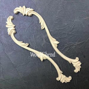 May include: Two cream-colored, curved wooden decorative appliques with a floral and scroll design. The appliques are arranged on a dark gray surface. The text "Wood Bend" is visible on the surface.