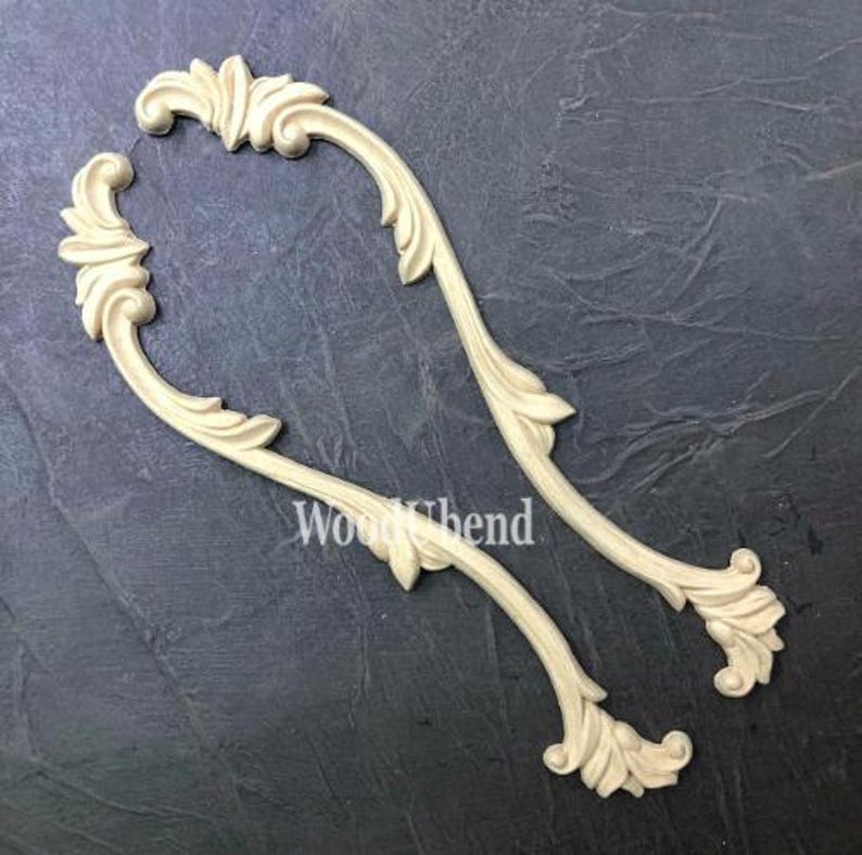 May include: Two cream-colored, curved wooden decorative appliques with a floral and scroll design. The appliques are arranged on a dark gray surface. The text "Wood Bend" is visible on the surface.
