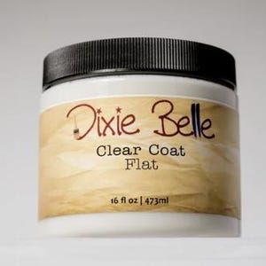 Może przedstawiać: Biały plastikowy słoik z czarną zakrętką, posiadający beżową etykietę z napisem Dixie Belle Clear Coat Flat oraz 473ml. Pojemnik zawiera przezroczysty płyn wykończeniowy, a na etykiecie znajduje się mała grafika przedstawiająca pędzel.